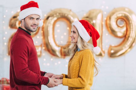 smiling couple in santa hats holding hands at home with 2019 new year golden balloons