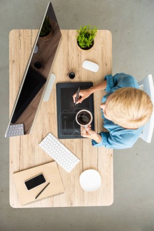 overhead view of female freelancer drinking coffee working on graphic tablet at table with smartphone and computer at home office 