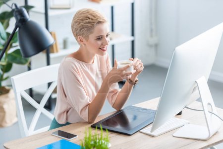 smiling female freelancer drinking coffee at table with graphic tablet and computer in home office 