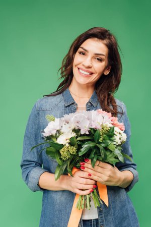 smiling attractive woman holding flower bouquet and looking at camera isolated on green