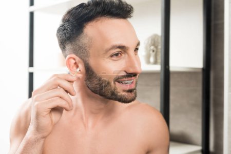cheerful shirtless man cleaning ear while looking at mirror in bathroom 