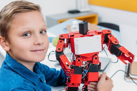 smiling schoolboy holding red robot and looking at camera in stem class