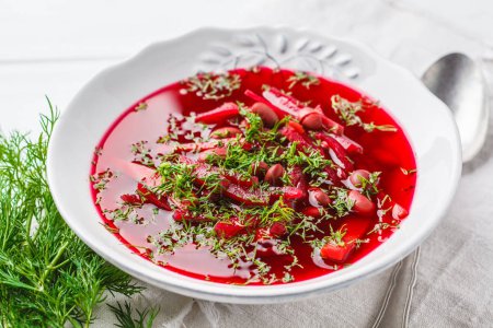 Vegetarian beet soup (borscht) with beans and vegetables in a white plate on a white wooden background.