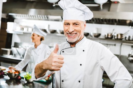 selective focus of handsome smiling male chef in uniform looking at camera and showing thumb up sign in restaurant kitchen