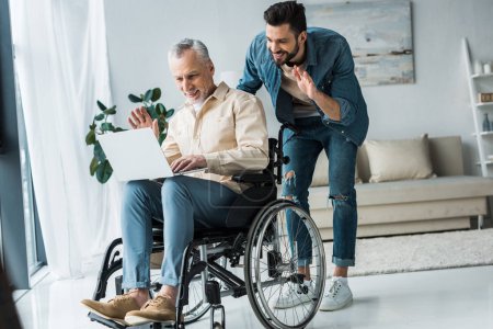 cheerful disabled retired man sitting in wheelchair near bearded son waving hand while having video call on laptop