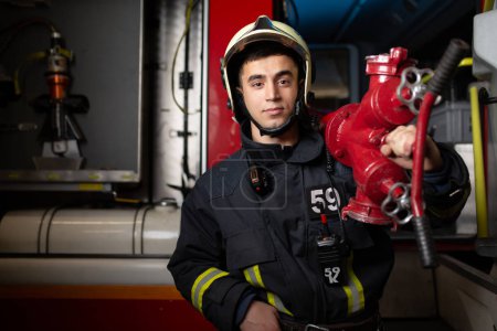 Photo of young man firefighter with fire cock on shoulder against background of fire truck