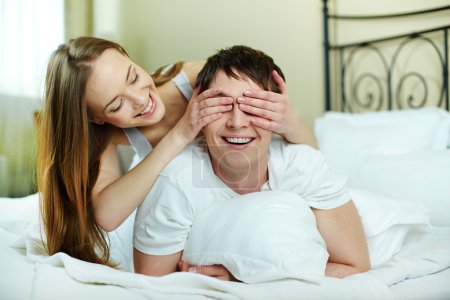 Happy girl closing her boyfriend eyes by hands in bed