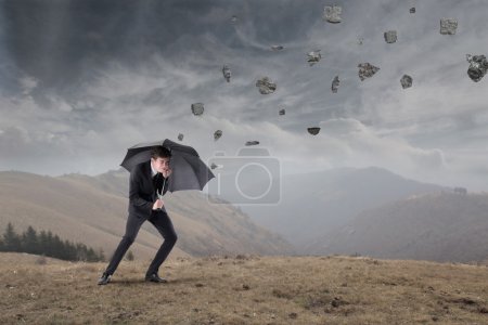 Businessman sheltering under an umbrella from rocks falling to the ground