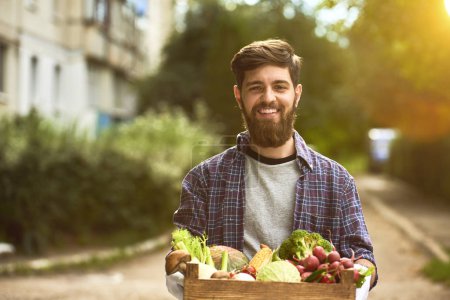 Young man holding wooden box with healthy organic vegetables, blurred summer park background