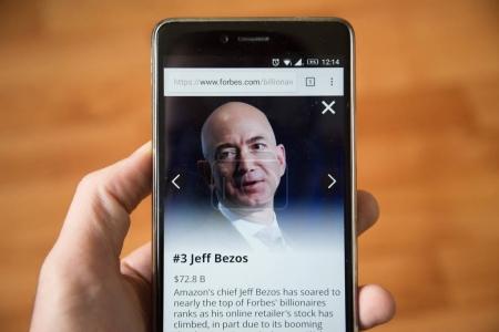 London, december 31, 2017: Jeff Bezos on forbes website, mobile phone version. Hand holding smartphone with wooden background