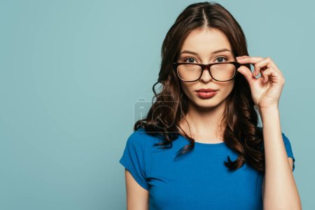 attractive, confident girl touching glasses while looking at camera isolated on blue