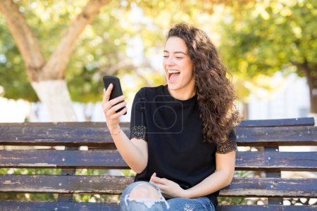 young excited woman sitting on a bench and making a video call with her smart phone