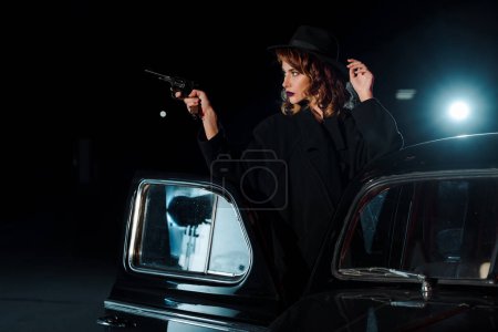 dangerous woman in hat holding gun near retro car on black 