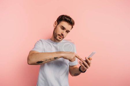 confident young man pointing with finger at smartphone during video call on pink background