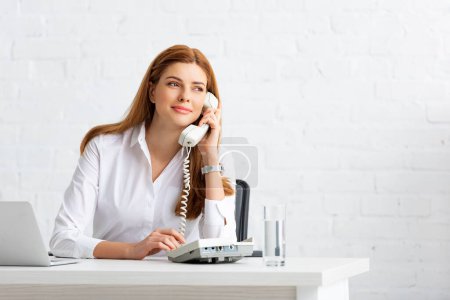 Smiling businesswoman looking away while talking on telephone at table in office