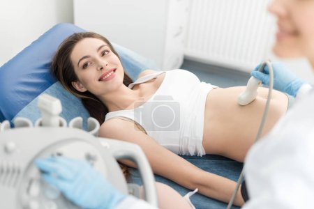 doctor examining belly of smiling pregnant woman with ultrasound scan in clinic
