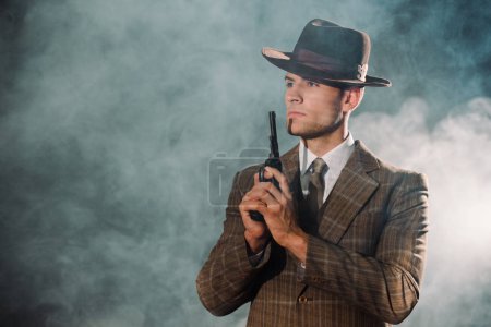 handsome gangster in hat holding gun on black with smoke 