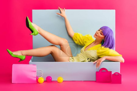 smiling woman in purple wig as doll lying in blue box with shopping bags and balls, on pink