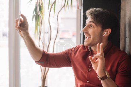 man in wireless earphones using having video chat on smartphone and showing peace sign near window