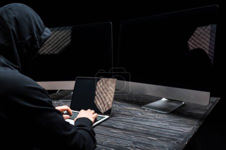 hacker using laptop with blank screen near computer monitors isolated on black 