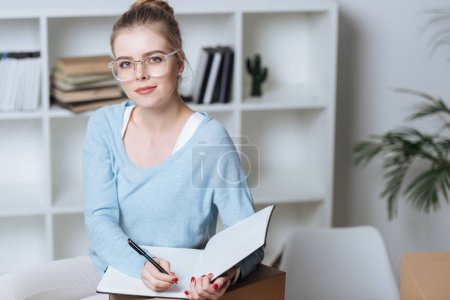 portrait of online shop proprietor with pen and notebook looking at camera at home office
