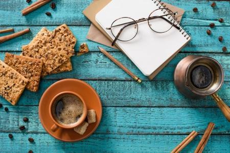 flat lay with cup of coffee, cookies, eyeglasses, empty notebook, roasted coffee beans and cinnamon sticks around on blue wooden tabletop