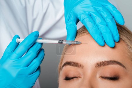 cropped view of beautician in latex gloves holding syringe near forehead of girl isolated on grey 