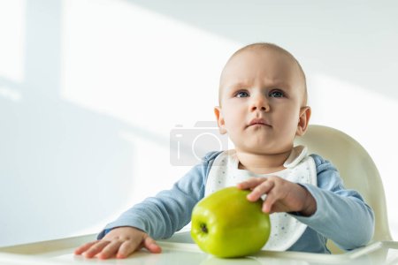 Cute baby boy touching green apple on table of feeding chair on white background