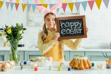 cute kid with bunny ears holding chalkboard with happy easter lettering and looking away