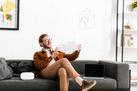 Smiling 3d artist showing peace symbol while having video call on digital tablet on couch in office 
