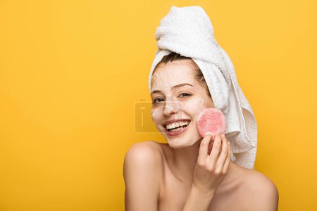 happy girl with moisturizing facial mask looking at cosmetic sponge isolated on yellow