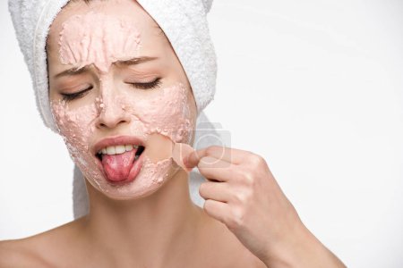 displeased girl removing peeling mask from face and sticking out tongue isolated on white