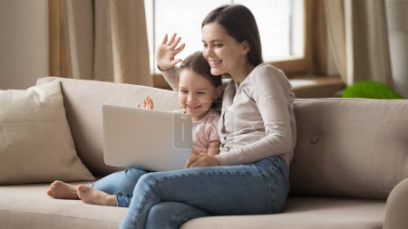 Smiling mom and little daughter talk on webcam using laptop